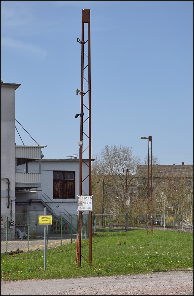 Anschlussverwendung der Oberleitungsmasten der badischen Staatsbahn in L�rrach-Brombach an einem ehemaligen Werksgleis. Kamerainstallation und Lampenmast. April 2013

Die ersten ernstzunehmenden Elektrifierzierungen mit Einphasenwechselstrom wurden anfangs mit 15 kV und 15 Hz durchgef�hrt. Durch einen Beschluss der badischen, bayrischen und hessisch-preu�ischen Staatsbahnen im Jahr 1912 wurden 16 2/3 Hz festgelegt. Die BLS schloss sich dem an und stellte ihre bereits elektrifizierte Strecke gleich darauf um, die RhB verwendete ebenso 16 2/3 aber eine reduzierte Spannung von 11 kV, die SBB hingegen begann erst ab 1919 mit Elektrifizierungen. In Deutschland waren Wiesen- und Wehratalbahn das gr��te elektrifizierte Netz ab 1913, immerhin bis 1936 mit 15 Hz. Der 1. Weltkrieg bremste erst einmal weitere Elektrifizierungen bis 1922 aus. Somit d�rften diese Masten 105 Jahre alt sein. Kurz vor Zell im Wiesental sind sogar noch einige dieser Masten in Betrieb. Im Bild der vierte Mast ist aus der Reichsbahnzeit.