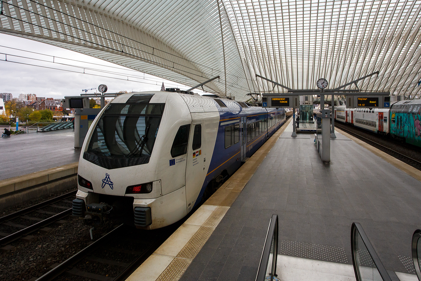 Der dreiteilige, 3-System Stadler FLIRT³ Triebzug 550 (94 84 4 040 550-x NL-AN) der Arriva Personenvervoer Nederland steht am 21 Oktober 2025 im Bahnhof Liège Guillemins (Lüttich-Guillemins), als Dreiländerzug / Drielandentrein / Train des trois pays bzw. RE18 / S 43 (LIMAX - Liège-Maastricht-Aachen-Express (Lüttich über Maastricht nach Aachen) zur Abfahrt bereit. 

Für den Einsatz in der Provinz Limburg beschaffte die Arriva Nederland 38 Triebzüge des Typs Stadler Flirt 3, die ab 2016 in Betrieb gingen. Zum Einsatz kommen dort 15 zwei- und 13 dreiteilige Fahrzeugkompositionen, sowie 8 dieser dreiteiligen Flirt´s in Mehrsystemausstattung (3-System) die von Arriva NL auf dem grenzüberschreitenden Dreiländerzug zwischen Lüttich über Maastricht nach Aachen eingesetzt werden. Die 8 Mehrsystem-FLIRT wurden 2017 bis 2020 in Dienst gesetzt. 

Im Februar 2016 bestellte Arriva Personenvervoer Nederland BV acht dreiteilige FLIRT-Elektrotriebzüge mit Niederflurtechnik bei Stadler. Die Dreisystemzüge können mit 1,5 kV Gleichstrom, 3 kV Gleichstrom und 15 kV Wechselstrom betrieben werden und verkehren im grenzüberschreitenden Regionalverkehr nach Belgien und Deutschland. Die neueste Generation der dreiteiligen FLIRT-Triebzüge ist für eine Höchstgeschwindigkeit von 160 km/h ausgelegt. Sie bieten 172 Sitzplätze, davon 12 in der ersten Klasse und 13 Klappsitze. Die Triebzüge verfügen über Luftfederung und breite Einstiegstüren. Dank des Niederfluranteils von 90 Prozent ist der Zugang zu den klimatisierten Zügen einfach, und die optimale Raumausnutzung gewährleistet ausreichend Platz für Kinderwagen und Rollstühle.

Der Dreiländerzug, niederländisch Drielandentrein, französisch train des trois pays oder LIMAX (Akronym aus Liège-Maastricht-Aachen-Express) ist eine Bahnverbindung, die von Arriva Personenvervoer Nederland betrieben wird.Sie verläuft von Aachen Hbf über Herzogenrath (Deutschland), Heerlen, Maastricht (Niederlande) und Visé nach Liège-Guillemins (Belgien).

Seit dem 29. Januar 2019 verkehren die Züge halbstündlich zwischen Maastricht und Heerlen sowie stündlich grenzüberschreitend bis Aachen. Inzwischen wurde der Halbstundentakt durch den zweigleisigen Ausbau zwischen Heerlen und Landgraaf bis Herzogenrath ausgeweitet. Seit Dezember 2023 wird ebenfalls Aachen zwei Mal pro Stunde erreicht, was vorher durch Trassenkonflikte mit dem RE 4 nicht möglich war. Die stündliche Verlängerung der Linie nach Lüttich fand am 30. Juni 2024 statt. Ziel ist, die Verbindung Lüttich–Maastricht–Aachen im 30-Minuten-Takt zu bedienen.

Der FLIRT (Akronym für Flinker Leichter Innovativer Regionaltriebzug) basiert auf dem Stadler GTW. Der Hauptunterschied besteht darin, dass die Antriebseinheiten des FLIRT hinter den Führerständen angeordnet sind (anstatt in einem separaten Triebwagen). Dieser Zugtyp zeichnet sich durch helle und geräumige Innenräume aus, die durch den Einsatz von Jacobs-Drehgestellen in Kombination mit breiten, offenen Übergängen und einem Niederfluraufbau ermöglicht werden. Der Niederfluraufbau, zusammen mit einer ausziehbaren Trittstufe, verbessert die Zugzugbenutzung für Menschen mit körperlichen Einschränkungen. Das geringe Gewicht des Zuges in Verbindung mit hohem Drehmoment macht ihn ideal für Züge mit häufigen und kurzen Haltezeiten.

Konstruktion:
Die FLIRT-Züge bestehen hier drei Wagen, die an den Wagenübergängen von Jacobs-Drehgestellen getragen werden. Der Einsatz von Jacobs-Drehgestellen ermöglicht kurze und breite Wagenübergänge und sorgt so für hohe Transparenz. Daher verfügen diese Züge über breite Wagenübergänge und einen Niederflureinstieg, wie er bei U-Bahnen und Straßenbahnen üblich ist. Die gute Sicht im gesamten Zug, kombiniert mit den Kameras, soll ein Gefühl der Sicherheit vermitteln. Die Bodenhöhe von 780 mm entspricht der der meisten Bahnsteige. Bis zu drei Züge können im Verbund verkehren.

Im Innenraum gibt es keine Trennwände und Türen; lediglich die erste und zweite Klasse sind durch Glastüren und Trennwände voneinander getrennt. Die Züge sind unter anderem mit Klimaanlage, einem Fahrgastinformationssystem mit TFT-Displays und automatischer Ansage, (Sicherheits-)Kameras und einem Notrufsystem ausgestattet, das nach Betätigung der Notbremse einen akustischen Kontakt zum Lokführer ermöglicht.

Interieur/Innenraum:
Fahrgäste haben Zugang zu USB-Ladestationen für mobile Geräte, Steckdosen und kostenlosem WLAN. Die Züge sind zudem mit einem Niederflurzugang, ausziehbaren Trittstufen und einer rollstuhlgerechten Toilette ausgestattet, um das Reisen für Menschen mit körperlichen Einschränkungen zu erleichtern. Diese Toilette verfügt über eine klappbare Wickelauflage. Die Züge bieten Abteile der ersten und zweiten Klasse, einen Salon und einen Fahrradabstellraum. Die Sitze der ersten Klasse (Kiel Match G2) sind grau mit orangefarbenen Kopfstützen. Die Sitze der zweiten Klasse sind blau-kariert mit dunkelblauen Kopfstützen.

Die 3-System-Züge:
Die Züge sind am neuen Arriva-Logo, den Aufklebern unterhalb der Führerstandsfenster mit dem Text „Kofinanziert von der Europäischen Union“ und den Dreiländerzug-Aufklebern erkennbar. Auf Wunsch Belgiens fehlt an den Führerstands Seiten der, sonst bei Arriva übliche, Limburg-Löwen. Die Züge sind primär für den Schnellzugverkehr konzipiert, sie verfügen über drei Türen pro Seite, viele Sitzplätze, wenige Klappsitze und wenige Fahrrad- oder Rollstuhlplätze. Im internationalen Verkehr werden Reiseinformationen auf Niederländisch, Deutsch und Französisch durchgesagt. Im Mittelteil gibt es zwei querangeordnete Sitzbänke. 

Die 3-System Stadler FLIRT³ (Baureihe IIImcs) erzeugen ein anderes Geräusch als die reine 1,5 kV- Gleichstrom-Triebzüge (Baureihe II und III). Dieses Geräusch wird durch die Traktionsumrichter verursacht, die es dem Triebzug ermöglichen, mit verschiedenen Spannungen zu arbeiten. Diese Umrichter sind von anderer Bauart und nutzen ebenfalls eine Drei-Phasen-Technologie, was ein leichtes Brummen des Triebzugs verursacht. Bei einer Oberleitungsspannung von 1,5 kV unterscheidet sich dieses Geräusch von dem bei 3 kV und 15 kV. Auch bei 1,5 kV ist ein Brummen hörbar, das von einer Drosselspule auf dem Dach stammt.

Technik der 3-System-Züge: 
Die 3-System-Triebzüge verfügen über zwei Stromabnehmer auf dem Mittelwagen, einen für Gleichstrom (1,5 und 3 kV) und einen für Wechselstrom (15 kV). Hinter jedem Führerstand befindet sich ein Stromumrichter (Fabrikat ABB, Type BORDLINE CC1500 MS_15-3-1.5kV_M_1300_070A01), der die Oberleitungsspannung in Drehstrom für die Fahrmotoren umwandelt. Die Umrichter arbeiten mit schnell schaltenden IGBTs; die Schaltfrequenz ist während der Fahrt hörbar. Beide Fahrmotoren (Fabrikat TSA, Type TMF 59-33-4 (600 kW)) eines Drehgestells werden gemeinsam gesteuert. Die Zugkraft der Fahrmotoren wird über ein Zahnradgetriebe mit einem Übersetzungsverhältnis von 1:5,347 auf die Achsen übertragen.

Auslieferung:
Der erste 3-System-Zug (550) wurde am 19. September 2017 über Venlo ausgeliefert. Da ein belgisches Gesetz (von 2014) die Ausstattung neuer Züge im Schienennetz mit dem ETCS-Sicherheitssystem vorschreibt, mussten die IIImcs entsprechend ausgerüstet werden. Dies erfolgte im Zeitraum 2021–2023 bei Stadler Refit in Blerick. Die ersten Testfahrten in Belgien fanden am 1. Juni 2022 statt, die Züge 550 und 555 waren bereits mit ETCS ausgestattet, andere noch nicht. Die endgültige Umrüstung auf ETCS erfolgte 2023, woraufhin Anfang 2024 Testfahrten und die Zulassung für Belgien folgten.

TECHNISCHE DATEN der 3-System Stadler FLIRT³:
Gebaute Anzahl: 8 (dreiteilig)
Nummerierung: 550 bis 557
Hersteller: Stadler Rail
Baujahre: ab 2017
Spurweite: 1.435 mm (Normalspur)
Achsfolge: Bo'2'2'Bo' (8 Achsen, davon 4 angetrieben) 
Länge über Kupplung: 63.200 mm
Fahrzeugbreite: 2.820 mm
Fahrzeughöhe: 4.120 mm
Drehzapfenlabstände: 17.600 mm / 17.500 mm / 17.500 mm / 17.600 mm
Achsabstand im Triebdrehgestell: 2.500 mm
Achsabstand im Laufdrehgestell: 2.700 mm
Treibraddurchmesser: 920 mm (neu)
Laufraddurchmesser: 760 mm (neu) / 690 mm (abgenutzt)
Leergewicht: 121 t
Höchstgeschwindigkeit: 160 km/h (in NL und B planmäßig 140 km/h) / 160 km/h (in D)
Sitzplätze: 12 in 1. Klasse, 147 der 2. Klasse davon 13 Klappsitze
Stehplätze: 161 (4 Personen/m²)
Bodenhöhe: Niederflur 780 mm (90%), Hochflur 1.180 mm
Türbreite: 1.300 mm
Türen je Längsseite: 3
Leistung am Rad: 3.000 kW
Anfahrzugkraft: 200 kN
Anzahl der Traktionsmotoren: 4 TSA Typ TMF 59-33-4 (600 kW)
Übersetzung: 1 : 5,347
Stromsysteme: 1,5 kV DC, 3,0 kV DC (Gleichstrom) und 15 kV 16,7 Hz AC (Wechselstrom)
Beschleunigung bis 54 km/h: 0,95 m/s²
Verzögerung: 1,15 m/s²
Kupplung: Scharfenberg