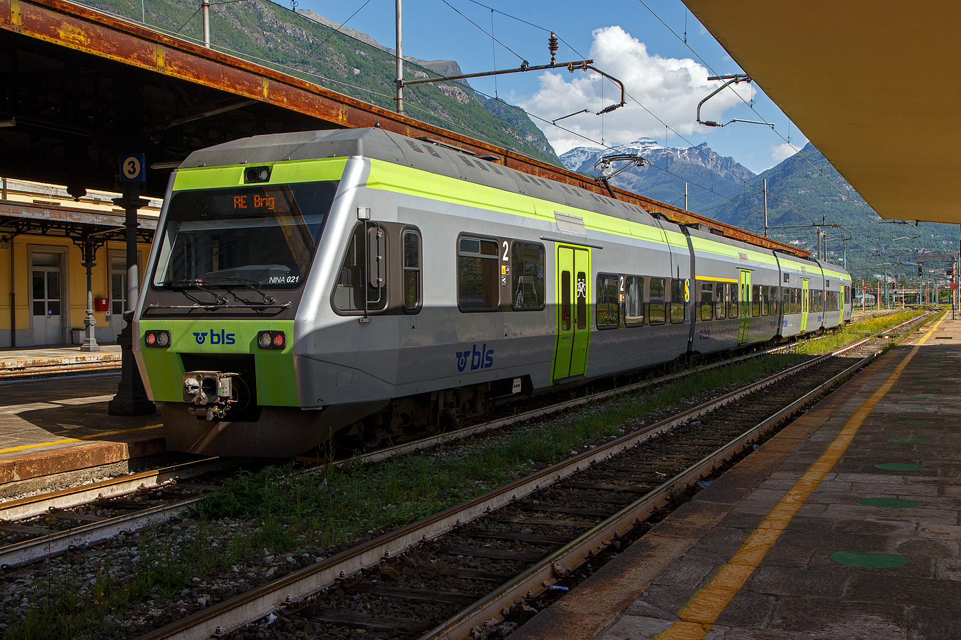 Der vierteilige BLS NINA 021 - RABe 525 021(94 85 7525 021-2 CH-BLS) steht am 26. Mai 2023 als RE nach Brig im Bahnhof Domodossola zur Abfahrt bereit.

Eigentlich sollten die BLS RABe 528 “MIKA” von Bern über die Lötschberg-Bergstrecke (Kandersteg) und Brig nach Domodossola fahren, aber diese haben noch keine Zulassung für Italien, die sie hinter dem SBB) Simplontunnel bis Domodossola brauchen. Und so wird der Zuglauf in Brig gebrochen und “NINA” fahren (min. bis zum Sommer) als Ersatz.

Von den BLS «NINA» (Niederflur-Nahverkehrszug) wurden 41 Stück zwischen 1998–2005 in 5 Serien von Vevey Technologies SA in Villeneuve (vormals ACMV - Ateliers de constructions mécaniques de Vevey) ab 2001Bombardier Transportation zusammen mit Bombardier-Talbot in Aachen (D)gebaut, die elektrische Ausrüstung wurde von Alstom/Traxis (vormals Holec) zugeliefert. An die zweite Bestellung der BLS schlossen sich auch die Transports de Martigny et Régions (TMR) und die Transports Régionaux Neuchâtelois (TRN) an, diese werden jedoch als RABe 527 geführt. 

Die klimatisierten Niederflur-Nahverkehrszüge „NINA“ bieten modernen Fahrkomfort in erfrischendem Design und verkehren sowohl als 3-teilig wie auch als 4-teilige Komposition. Tiefe Einstiege und große Plattformen sorgen für Bewegungsfreiheit, und Geschwindigkeiten von bis zu 140 km/h garantieren attraktive Fahrzeiten. Die dreiteiligen bieten 
148 und die vierteiligen 191 Sitzplätzen. Es können bis zu 4 Triebzüge aneinandergekoppelt werden (maximal 592 bis 764 Sitzplätze).

Das Fahrzeug wurde Mitte der 1990er-Jahre von Vevey Technologies zusammen mit Bombardier-Talbot in Aachen (D), entwickelt und gebaut. Das Niederflurkonzept und die Drehgestell-Technik stammen weitgehend vom Triebzug Talent, der von Bombardier-Talbot in Aachen entwickelt wurde. Die Drehgestelle weisen eine fast gleiche Konstruktion auf, die lediglich für die NINA angepasst wurde. Für die Elektrotechnik war die Firma Holec Machines & Apparaten – die spätere Alstom/Traxis – aus Ridderkerk (NL) zuständig, die, wie die Vevey Technologies zu dieser Zeit auch, zur Begemann-Gruppe aus den Niederlanden gehörte. Seit 2001 firmiert das Unternehmen Vevey Technologies als Bombardier Transportation (Schweiz). Die Fahrzeuge wurden weiterhin im Werk in Villeneuve VD hergestellt.

Die «NINA» wurden als zwei- bis vierteilige Triebzüge angeboten. Angetrieben werden sie von zwei zweiachsigen Triebdrehgestellen unter den Führerständen. Zwischen den einzelnen Wagen sind Jakobsdrehgestelle angebracht. Der Zug zählt wie der Stadler GTW zu den Leichtbaufahrzeugen. Eine dreiteilige Einheit wiegt leer nur 78 Tonnen (zum Vergleich eine Re 465 Lok  wiegt 84 Tonnen). Die Höchstgeschwindigkeit beträgt 140 km/h.

Die BLS beschaffte 1998 eine erste Serie von acht dreiteiligen Einheiten. Diese Fahrzeuge wurden nachträglich mit einem WC ausgerüstet. Im Jahre 2000 folgte eine Nachbestellung über sechs Fahrzeuge, denen eine dritte Serie über 18 Fahrzeuge folgte, die Ende 2002 bestellt wurden. Mit der im Jahre 2003 erfolgten vierten Bestellung über vier Fahrzeuge erhöhte sich der Bestand auf insgesamt 36 Einheiten. Um die Sitzplatzkapazität zu erhöhen, wurden im Jahre 2007 die dreizehn RABe 525 015 bis 027 um je einen Zwischenwagen zu vierteiligen Triebzügen verlängert. Ab 2009 erhielten die NINA eine Klimaanlage.

An die zweite Bestellung der BLS schlossen sich auch die Transports de Martigny et Régions (TMR) und die Transports Régionaux Neuchâtelois (TRN) an. 

TECHNISCHE DATEN:
der RABe 525 NINA dreiteilig nach Refit, in Klammern vierteilig
Spurweite: 	1.435 mm (Normalspur)
Achsfolge: 	Bo'2'2'Bo'   (Bo'2'2'2'Bo')
Länge über Puffer: 47.740 mm  (61.900 mm)
Achsabstand im Triebdrehgestell: 2.500 mm
Achsabstand im Laufdrehgestell (Jakobsdrehgestell): 2.700 mm
Höhe: 4.315 mm
Breite: 3.030 mm
Gewicht: 79,0 t  (96.0 t)
Dauerleistung: 1.000 kW / 1.360 PS
Anfahrzugkraft: 105 kN
Höchstgeschwindigkeit: 140 km/h
Stromsystem: 15 kV 16⅔ Hz AC
Sitzplätze in der 1. Klasse: 20  (20)
Sitzplätze in der 2. Klasse: 114  (159)
Rollstuhlplätze: 1  (1)
Fahrradplätze/Veloplätze: 3  (9)

Quellen: BLS, wikipedia
