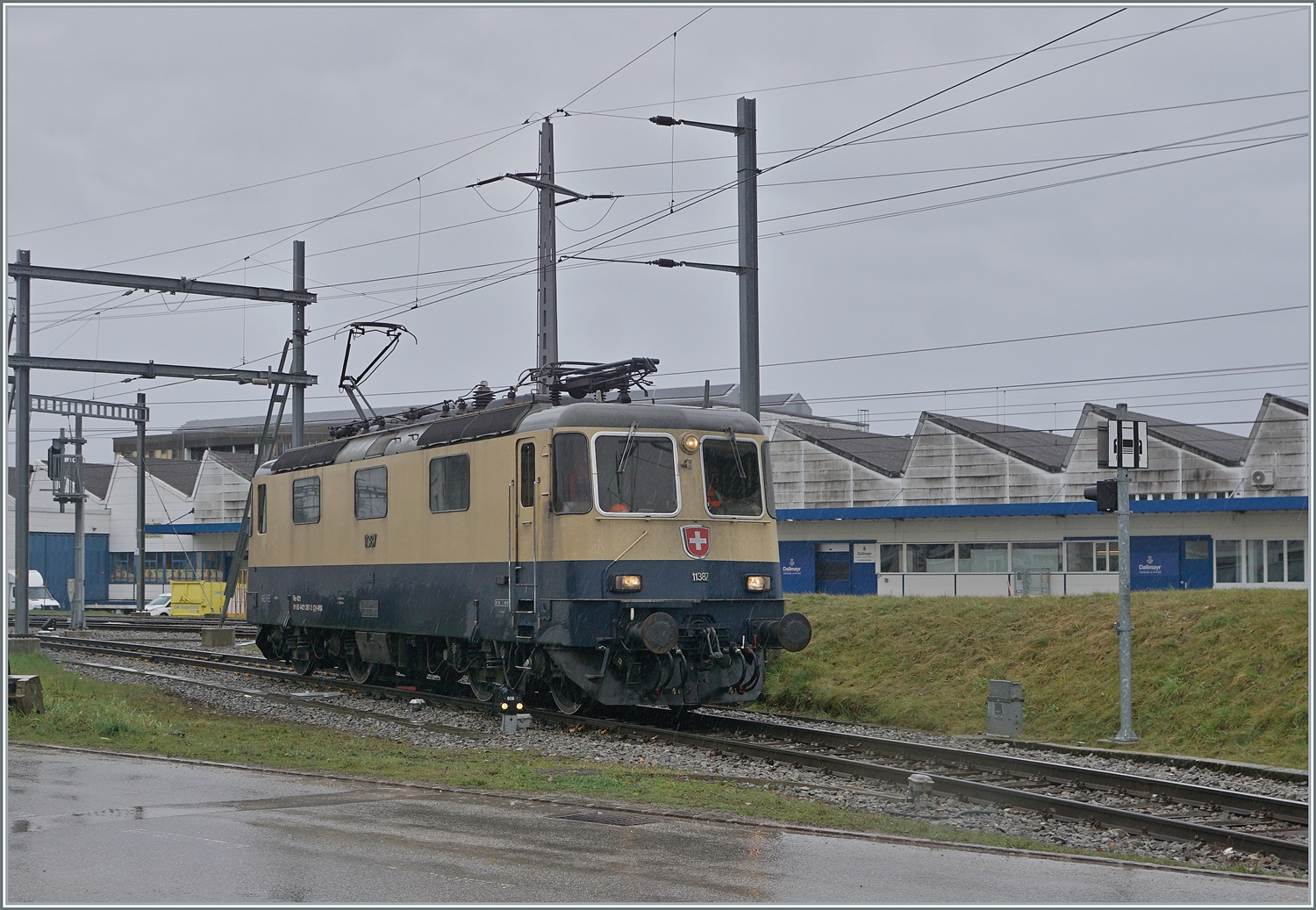 Die von Morges gekommene IRSI Re 4/4 11387 verlässt in Gland die Strecke nach Genève und fährt in über ein Industriegleis in Richtung Kieswerk um dort ihren Zug nach Apples abzuholen. Die angemietete IRSI/IGE Re 4/4 II 11387 (Re 421 387-2) ersetzt die zur Zeit in der Revision in Bellinzona weilende BAM MBC MBC Re 420 506.

22. Feb. 2024