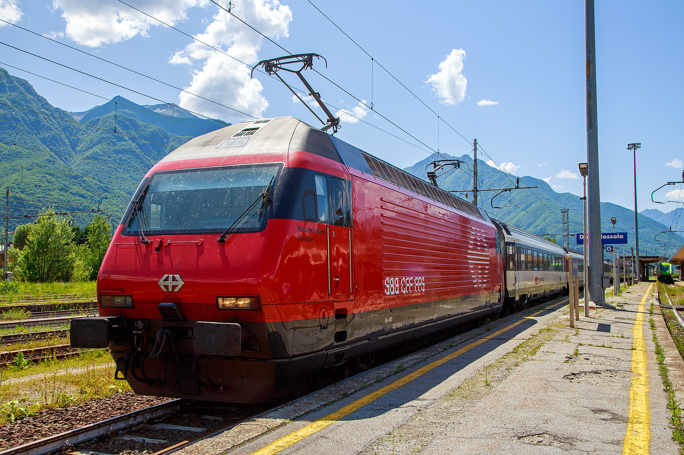 Die SBB Re 460 016-0  Rohrdorferberg Reusstal   (91 85 4 460 016-0 CH-SBB) hat am 26 Mai 2023 mit dem IR 3017 (Brig – Domodossola) den Zielbahnhof Domodossola (I) erreicht und ist nun mit dem Zug auf Gleis 6 abgestellt. Nach gut 2 ½ Stunden (um 13:48 Uhr) geht es dann als IR3022 von Domodossola zurück nach Brig.

Die Lok2000 wurde 1992 von der SLM (Schweizerischen Lokomotiv- und Maschinenfabrik in Winterthur) unter der Fabriknummer 5477 gebaut, der elektrische Teil ist von der ABB (Asea Brown Boveri). Sie war 2015 der 2. Prototyp der im SBB Werk Yverdon-les-Bains (VD) das vollständige Modernisierungs-/Refit-Programm LOK2000 durchlief. So sollen die Loks fit für die nächsten 20 Jahre sein, zudem werden durch alle 119 SBB Re 460 Lokomotiven so etwa 30 Gigawattstunden Energie eingespart.

Modernisierungs-/Refit-Programm der LOK2000 (Re 460):
Zwischen 2015 und November 2022 unterzog die SBB alle ihrer 119 Re 460 Lokomotiven (LOK2000) einem Refit-Programm (Modernisierung) im SBB Werk Yverdon-les-Bains (VD). Dabei wurden unter anderem die auf dem GTO-(Thyristor)-Halbleiter-Technologiestand der 1990er-Jahre basierende Leistungselektronik (GTO- Traktionsumrichter) durch IGBT-Traktionsumrichter (insulated-gate bipolar transistor) der neuesten Generation von ABB ersetzt, welche die Fahrmotoren mit Strom speisen und Bremsenergie zurückgewinnen. Die SBB hat damit, in Zusammenarbeit mit der ABB, die gesamte Flotte Re 460 modernisiert und hat diese auf den neuesten Stand der Antriebstechnik gebracht. Die gesamte Flotte spart dank neuen Traktionsumrichtern und weiteren technischen Optimierungen jedes Jahr rund 30 Gigawattstunden Energie ein, das sind fünf Gigawattstunden mehr als ursprünglich erwartet. Die Energieeinsparungen entsprechen dem durchschnittlichen Stromverbrauch von 10.000 Schweizer Haushalten. Die Umrüstung auf modernste IGBT-Technologie und mit Wasser statt Öl betriebene Kühlsysteme der Umrichter ermöglicht eine Senkung des Energieverbrauchs und sorgt für einen ökologischeren und sicheren Bahnbetrieb.

Geschichte und Beschreibung:
Die SBB bestellte im Zusammenhang mit den Projekten Bahn 2000 eine erste Serie von 12 Universal-Hochgeschwindigkeitslokomotiven, noch als Re 4/4VI – 10701 bis 10712 bestellt. Diese Bestellung wurde noch vor Beginn der Konstruktionsarbeiten auf 24 erhöht. Für den alpenquerenden Güterverkehr wurde von der Eidgenossenschaft eine weitere Serie von 75 Lokomotiven (auch als 'Hupac'-Lokomotiven bekannt) in Auftrag gegeben. Die vorgegebenen Liefertermine für dieses Los waren derart kurz, dass die SLM an die Kapazitätsgrenze ihrer Werkstätte gelangte. Es wurden deshalb dreißig Lokkasten an Krauss-Maffei in München untervergeben 

Im Hinblick auf die geplante Hochgeschwindigkeitsstrecke wurden nochmals zwanzig Lokomotiven nachbestellt, was die Serie auf 119 ansteigen ließ. Die im Volksmund Lok 2000 genannte Lokomotive hat eine Höchstgeschwindigkeit von 230 km/h und eine Leistung von 6,1 MW und ist für den Schnellzugs- wie Güterverkehr ausgelegt. Um die Gesamtmasse von 84 Tonnen nicht zu überschreiten musste ein gesickter Leichtbaukasten konstruiert werden, dessen Form vom Designstudio Pininfarina entworfen wurde.

Um auf den Strecken durch die Alpen den Radverschleiß gering zu halten, haben die Lokomotive radial einstellbare Achsen erhalten. Auch auf dem Gebiet der elektrischen Ausrüstung wurde Neuland beschritten. Zwei Ebenen Steuerelektronik für das Fahrzeug und den Antrieb sind eingebaut. Über das Fahrzeugleitgerät wurden zu viele Elemente gesteuert, wie Scheibenwischer oder Signalhorn. Dies führte anfänglich zu vielen Störungen und oft blieben Lokomotiven auf der Strecke liegen. Erst die Verbesserung der Programmierung erlaubte es die Zuverlässigkeit so zu erhöhen, dass es kaum mehr zu Ausfällen führte. Als Feststellbremse wurden in den Drehgestellen Permanentmagnetschienenbremsen eingebaut. Auch hier gab es am Anfang übermäßig viele Störungen und die Lokomotiven klebten regelmäßig an den Schienen fest.

Um den Schiebedienst auf den Gebirgsstrecken weniger personalaufwendig durchzuführen, wurden mehrere Lokomotiven mit einer amerikanischen Funkfernsteuerung ausgerüstet und als Ref 460 bezeichnet. Damit konnte von der Zugspitze aus eine am Zugschluss eingesetzte Lokomotive ferngesteuert werden. Die Divisionalisierung der SBB erlaubte es nicht mehr groß davon zu profitieren, denn alle Lokomotiven wurden dem Personenverkehr zugeteilt und der Einsatz vor Güterzügen war somit beendet.

Die Re 460 blieb nicht ein Einzelstück und entwickelte sich zu einer Lokomotivfamilie. Eine erste Bestellung wurde von der BLS für acht Lokomotiven gemacht. Die BLS Re 465 hat im Gegensatz zur Re 460 vier statt zwei Stromrichter. Jeder Motor hat dadurch seine eigene Regelung um die Eigenschaften im Bergeinsatz zu verbessern. Weiter können die BLS-Lokomotiven mit allen gängigen Schweizer Lokomotiven in Vielfachsteuerung verkehren. Das erlaubte es bei der BLS den Lokomotiveinsatz flexibler zu gestalten. Die SBB bestellte ebenfalls zehn Re 465, die bei Ablieferung schon das blaue BLS-Design hatten, aber erst später von der BLS übernommen wurden. Nach Finnland konnten auch in zwei Serien Lok 2000 verkauft werden. Dort sind sie als Sr2 bezeichnet. Nach Versuchsfahrten in Norwegen im schwierigen Winterdienst bestellten die NSB die fast baugleichen Lokomotive EL18 2241 bis 2262. Eine letzte Lieferung umfasst zwei Lokomotiven für Hongkong. Sie werden an jedem Ende eines Doppelstockzuges eingesetzt. Dies sind die letzten von SLM in Winterthur gebauten Fahrzeuge. Die Re 460 ist bis auf weiteres die letzte Vollbahn-Streckenlokomotive, die komplett von schweizerischen Unternehmen konstruiert und gebaut wurde. Der Lokomotivbau in der Schweiz wurde inzwischen aus wirtschaftlichen Gründen eingestellt. Einzig Stadler Rail stellt in der Schweiz noch Lokomotiven für den Rangier- und für den Zahnradbetrieb her.

TECHNISCHE DATEN der Re 460:
Nummerierung: 	Re 460 000–118
Anzahl: 119
Baujahre: 1991–1996
Hersteller: SLM - Winterthur / ABB (Asea Brown Boveri)
Design: Pininfarina
Hersteller Lokkasten: teilweise (30 Stück) Krauss-Maffei (München)
Spurweite: 1.435 mm (Normalspur)
Achsformel: Bo'Bo'
Länge über Puffer: 18.500 mm
Höhe: 4.300 mm
Breite: 3.000 mm
Drehzapfenabstand: 11.000 mm
Achsabstand im Drehgestell:  2.800 mm
Laufraddurchmesser: 1.110 mm (neu)
Dienstgewicht: 84,0 t
Höchstgeschwindigkeit:  200 km/h (zugelassen)  / 230 km/h (Techn. möglich) 
Stundenleistung: 6.100 kW / 8.300 PS
Dauerleistung: 4.800 kW / 6.000 PS
Anfahrzugkraft: 300 kN
Dauerzugkraft: 275kN
Stromsystem: 15 kV, 16,7 Hz (über Oberleitung)
